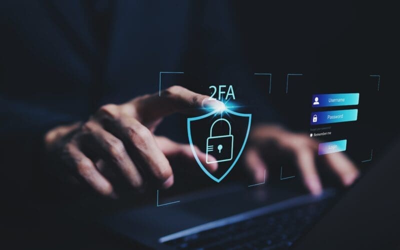Człowiek trzymający ręce na klawiaturze laptopa i hologramy symbolizujące bezpieczeństwo oraz system 2FA